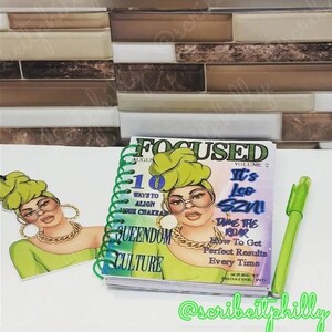 May include: A green and black spiral bound journal with the title "Focused" and the text "It's Leo SZN!" on the cover. The cover also features a cartoon illustration of a woman with green hair and glasses. The journal is on a table with a green pen.