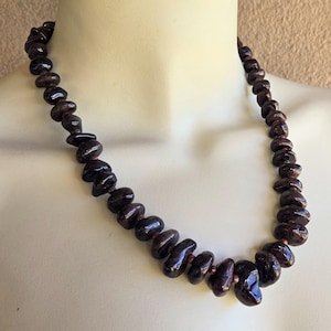 May include: A dark burgundy necklace composed of irregularly shaped beads. The beads are strung together, creating a statement piece. The necklace is displayed on a mannequin, showcasing its length and design. The beads have a polished, glossy finish.