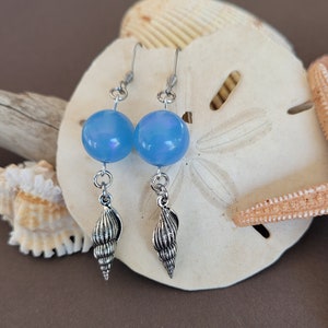 May include: A pair of blue iridescent glass bead earrings with silver shell charms. The earrings are displayed on a seashell and starfish.