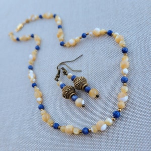 May include: A necklace and earrings set made with beige and blue beads. The necklace is made with round, flat, and oval-shaped beads. The earrings are made with round beads and a gold-colored accent.