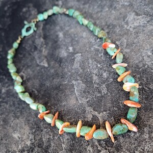 May include: A turquoise and orange stone necklace with a gold clasp. The necklace is made of irregular shaped stones and is strung on a thin gold chain.