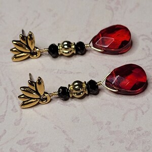 May include: A pair of dangle earrings with red teardrop-shaped glass beads, black beads, and gold accents. The earrings feature a gold flower design at the top.