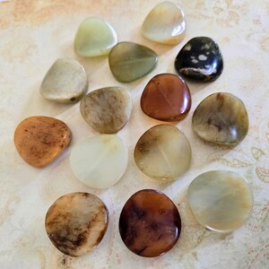 May include: A collection of 14 smooth, polished, natural stone coasters in various shades of brown, beige, and green. The coasters are shaped like rounded triangles and have a natural, organic look.
