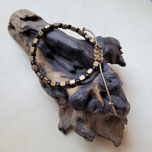 May include: A black and gold beaded bracelet with a knotted leather closure. The bracelet is displayed on a piece of driftwood.