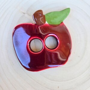 May include: A glossy, red ceramic apple-shaped object with two circular holes. It features a brown stem and a green leaf. The apple is set against a light wood grain background, creating a contrast of colors and textures.