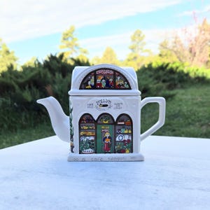 May include: White ceramic teapot with a cafe design. The teapot features a rectangular shape with a spout, handle, and lid. The front of the teapot depicts a cafe scene with the text "Polly's Home Made Teapot Co. Sweets and Cakes".
