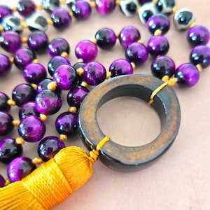 May include: A close-up of a beaded necklace with purple and black beads. The necklace has a large, dark wooden ring and a yellow tassel. The beads are strung on a yellow cord.