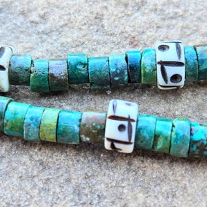 May include: Close-up of two strands of turquoise and white beaded jewelry. The turquoise beads are cylindrical and have a mottled green and blue appearance. The white beads are rectangular with black etched designs. The jewelry is displayed on a textured, light gray surface.