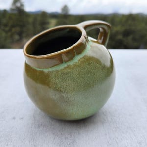 May include: A handcrafted, round ceramic mug with a handle. The mug features a glossy, speckled green glaze with a darker green rim and handle. The mug is designed for beverages.
