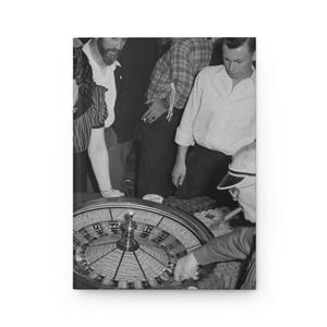 Könnte beinhalten: Schwarz-Weiß-Fotografie eines Rouletterads in einem Casino, umgeben von Menschen. Das Rad dreht sich, und die Kugel ist kurz davor, zu landen. Menschen beobachten das Spiel.