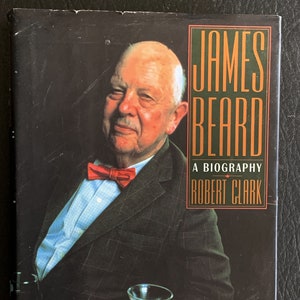 May include: A book cover featuring a portrait of James Beard, a famous American chef and cookbook author. The cover is black with a green and orange title block that reads "JAMES BEARD A BIOGRAPHY ROBERT CLARK".