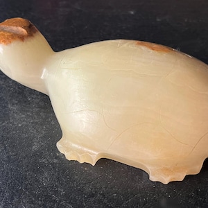 May include: A carved stone turtle figurine in shades of cream and brown. The turtle has a smooth, polished body with a textured shell. The head and tail are a darker brown color. The figurine is a decorative item.