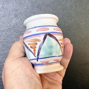 May include: A small, white ceramic vase featuring a hand-painted design with horizontal blue stripes, pink dashes, and triangular motifs in shades of teal, dark purple, and orange. The vase has a rounded body, a flared rim, and a pedestal base.