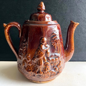 May include: A glossy brown ceramic teapot featuring a raised relief design of a figure standing near a well and trees. The teapot has a curved handle, a pointed spout, and a matching lid with a small knob handle on top, all in a uniform brown glaze.