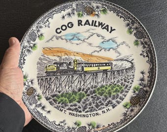 May include: A decorative plate featuring a yellow and black train on a trestle bridge, with the text "COG RAILWAY" and "MT. WASHINGTON, N.H." The plate has a detailed border with pine cone and foliage designs.