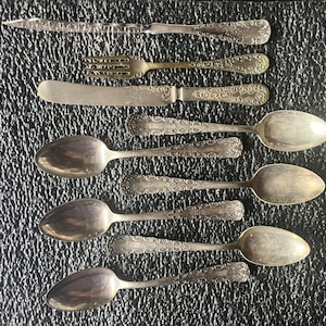 May include: An assortment of antique silverware, including spoons, a fork, and a knife, arranged on a textured black surface. The silverware features ornate, engraved designs and a tarnished silver finish, showcasing a vintage aesthetic.