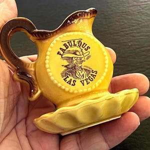 May include: A small yellow ceramic creamer pitcher with a brown handle and rim. The front features a circular emblem with a brown illustration of a cowboy and the text "FABULOUS LAS VEGAS" printed in brown. The base has a scalloped edge design.