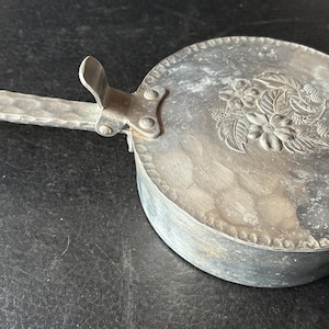 May include: A small, antique silver-colored container with a handle and lid. The lid features a floral design. The handle and body have a hammered texture. The container is likely made of metal.