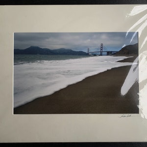 Puede incluir: Fotografía enmarcada del Golden Gate Bridge en San Francisco. La imagen muestra el puente a lo lejos, con una playa y el océano en primer plano. El cielo está nublado y el agua tiene olas blancas y espumosas.