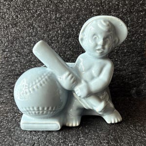May include: A light blue ceramic figurine of a child wearing a hat, holding a baseball bat and leaning against a baseball. The figurine is a decorative item, likely for a shelf or display.