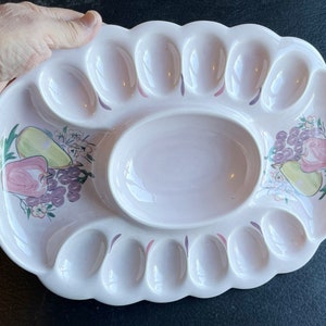 Peut inclure: Un plat à œufs en céramique blanche avec un grand creux central et 12 dépressions individuelles en forme d'œuf. Le plat est décoré d'un motif floral rose et violet et d'une poire.