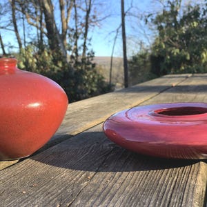 May include: Two red ceramic vases sit on a wooden surface. One is a rounded, bulbous shape with a narrow neck. The other is a flat, circular dish with a central hole and three small feet. Both have a glossy finish.