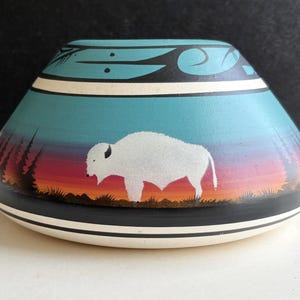 May include: Hand-painted ceramic vase with a Native American design. The vase features a white bison against a sunset backdrop of orange, red, and purple hues. The top has a turquoise background with black and white accents.