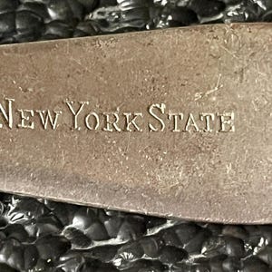 May include: Close-up of a tarnished metal object with the words "New York State" engraved in capital letters. The object has a rectangular shape with rounded edges and a textured surface. The background is dark and out of focus.