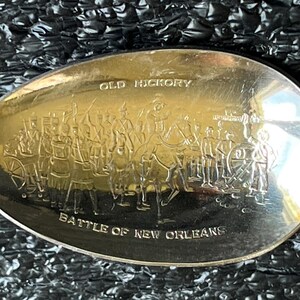 May include: Silver spoon with an engraved scene depicting a historical battle. The spoon bowl features the words "OLD HICKORY" and "BATTLE OF NEW ORLEANS" above and below a detailed illustration of soldiers and horses.
