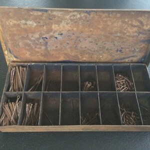May include: A vintage metal box, showing signs of rust, with a hinged lid and multiple compartments. The box is filled with an assortment of small, rusty metal pins and fasteners, suitable for various craft or hardware uses.