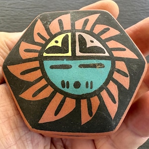 May include: A hexagonal terracotta tile featuring a stylized sun face design. The sun has a turquoise lower face, a black background, and radiating orange petals. The upper portion includes yellow and light pink geometric eye shapes.