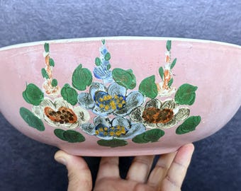 May include: A round ceramic bowl with a soft pink exterior finish. The bowl features a hand-painted floral design consisting of three clusters of flowers in shades of blue, white, and orange, accented with green leaves and tall vertical stems.