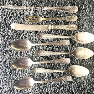May include: An assortment of antique silverware, including spoons, forks, and knives, arranged on a textured dark surface. The silverware features ornate, detailed patterns on the handles and blades, showcasing a vintage aesthetic. The fork has a gold color.