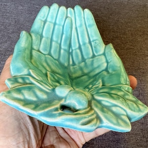 May include: A glossy, light turquoise ceramic dish shaped like two cupped hands holding a cluster of leaves and small round berries at the base. The surface has a smooth, reflective glaze with visible finger and leaf details molded into the clay.