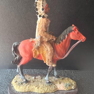 May include: A detailed figurine of a Native American chief riding a red horse. The chief wears a feathered headdress and traditional clothing. The horse has a black mane and tail, and the base is a brown wooden oval.