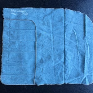 May include: A folded, light blue cloth with a rectangular shape and rounded corners. The cloth has horizontal stitched lines and the text "Sykes-Libby Goods" in the upper left corner. The edges are finished with white stitching.