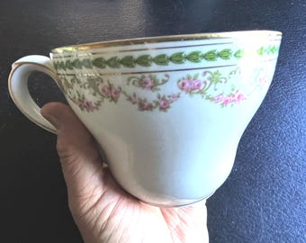 May include: A white porcelain teacup featuring a delicate gold rim and a decorative border of green laurel leaves. The upper section is adorned with a repeating pattern of small pink rose clusters and light green scrollwork accents.