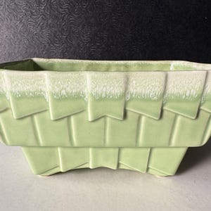 May include: A rectangular, light green ceramic planter with a woven basket design. The top edge features a textured, white trim. The planter is suitable for indoor or outdoor use and is a decorative item for plants.