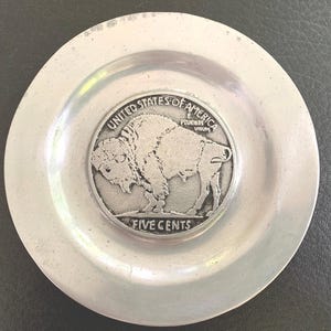 May include: A silver-colored ashtray with a recessed center. The center features a detailed image of a buffalo and the words "FIVE CENTS" and "UNITED STATES OF AMERICA".