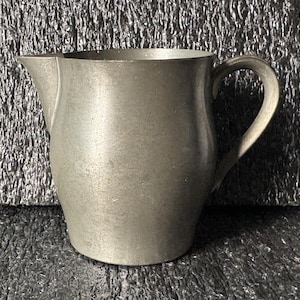 May include: A pewter pitcher with a curved handle and a spout for pouring. The pitcher has a smooth, metallic surface and a slightly rounded body. The overall design is simple and classic, suitable for serving beverages.
