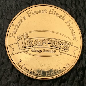 May include: Gold-colored metal coin with the words "Parker's Finest Steak House" and "Trapper's Chop House" on it. The coin also says "Limited Edition".