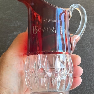 May include: A red glass pitcher with a clear glass handle and a diamond-patterned base. The pitcher is etched with the words "Souvenir Boone, Ia."