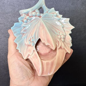 May include: A ceramic wall pocket shaped like a large grape leaf with a glossy finish. The piece features a gradient of soft pastel pink and light blue colors, with raised details of small grape clusters and textured veins across the leaf surface.