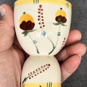 May include: A cream-colored ceramic egg cup featuring a crackled glaze finish. The exterior is decorated with hand-painted yellow and brown floral motifs, green stems, and dotted patterns. A yellow band encircles the top and bottom edges.