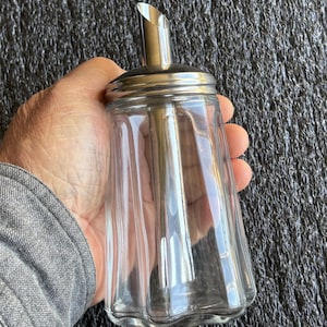 May include: A clear glass sugar dispenser with a stainless steel lid and spout. The dispenser has a fluted design and is held in a hand. The spout is angled for easy pouring. A classic kitchenware item.
