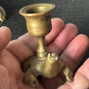 May include: A small, antique brass candlestick holder. The candlestick has a turtle base with a cup-shaped top. The brass has a warm, golden hue. The candlestick is designed to hold a single candle.