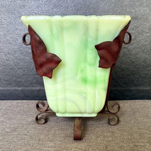 May include: A pale green marbled glass vase with a square, fluted body. The vase is held within a dark brown metal frame featuring decorative leaf-shaped accents on the sides and scrolled feet at the base, providing a stable support structure.
