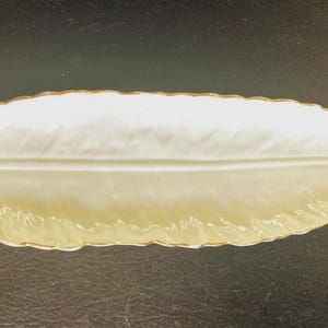May include: A long, narrow, leaf-shaped ceramic dish in an off-white color with a glossy finish. The edges of the dish are textured to resemble a serrated leaf, and the rim is accented with a thin gold-colored trim. A small circular hole is present.