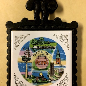 May include: A black metal trivet with a decorative handle and a square tile insert. The tile features a colorful illustration of Boston landmarks, including the U.S. Constitution and the Old North Church, with the words "Boston Mass Beans".