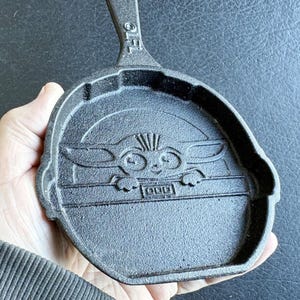 May include: Black cast iron skillet with a handle. The skillet features a raised relief of a character with large ears and eyes. The handle has the letters "OFL" embossed on it. The skillet is held in a hand.
